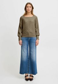 Olivgrüner Strickpullover mit Kabelmuster, kombiniert mit weit geschnittenen blauen Jeans. Die Textur ist weich; die Hose hat eine allmähliche Ausbleichung.