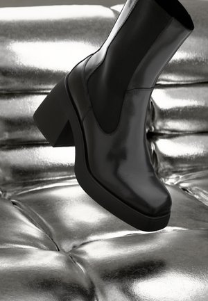 Glanzende zwarte leren enkellaars met chunky hak rustend op een metallic, doorgestikte zilveren kussen.