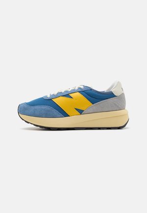 Baskets bleues et grises avec logo latéral jaune, lacets blancs, semelle épaisse beige et semelle noire sur fond blanc.