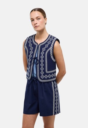 Jeune femme aux cheveux attachés en arrière portant un gilet sans manches bleu marine avec broderies géométriques blanches et un short bleu marine assorti avec broderie latérale.