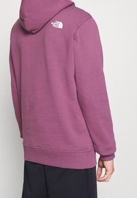 Lila fleece-tröja med dragkedja i huven. Har en vit logotyp på baksidan samt ribbade ärmslut och nertill. Strukturerad tyg.