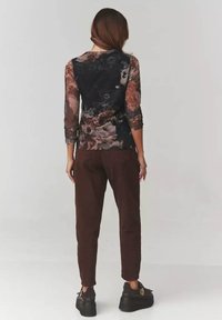 Kobieta zwrócona plecami, ubrana w długi rękaw, ciemną bluzkę w kwiatowy wzór, brązowe spodnie i czarne buty na platformie, stojąca na jednolitym tle.