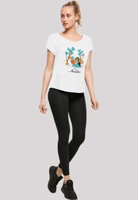Hvit kortermet t-skjorte med et fargerikt "Aladdin"-grafikk som viser karakterer og palmer, kombinert med svarte leggings og svarte joggesko.