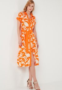 Orangenes Blumenkleid mit kurzen Ärmeln, tailliertem Schnitt, Knopfverschluss und einem seitlichen Schlitz. Hergestellt aus glattem Stoff mit weißen Blumenmustern.