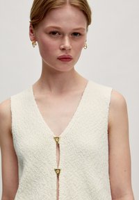 Cremfarbener, strukturierter Weste mit tiefem V-Ausschnitt und zwei dreieckigen goldenen Verschlüssen an der Vorderseite. Einfaches und minimalistisches Design.