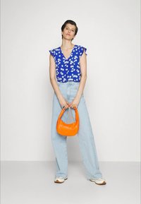 Blusa azul com padrões florais e mangas curtas com babados, combinada com jeans claros. Acessórios incluem uma bolsa laranja vibrante. Ténis brancos completam o visual.