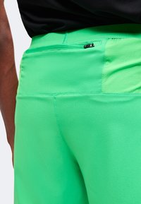 Gros plan sur un pantalon de sport vert vif avec une petite poche zippée sur la ceinture arrière, porté avec un haut noir.