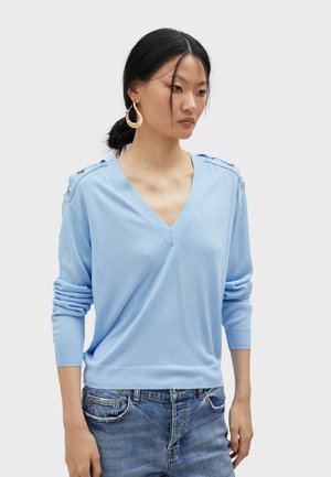 Lichtblauwe gebreide trui met een V-hals, lange mouwen en schouderknopen. Draagt met denim jeans. Gladde textuur.