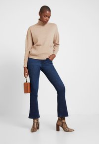 Bež pulover iz pletenega materiala z visokim ovratnikom in sprednjimi žepi, kombiniran s temno modrimi razširjenimi kavbojkami in rjavimi gležnjarji z vzorcem kačje kože. Rjava okrogla torbica.