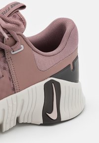 Rosa treningssko med teksturert mesh-overdel, avrundet form, hvit yttersåle og svarte detaljer. Har en fremtredende logo på helen.