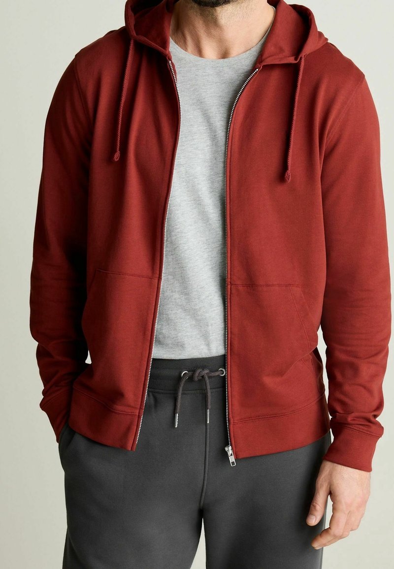 Sudadera roja con cremallera y capucha ajustable, hecha de un tejido suave. Llevada sobre una camiseta gris claro, combinada con pantalones de chándal gris oscuro.