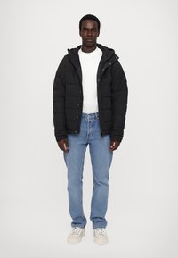Black padded jacket with a front zip, white t-shirt, blue jeans, and light sneakers. Model standing against a plain background.