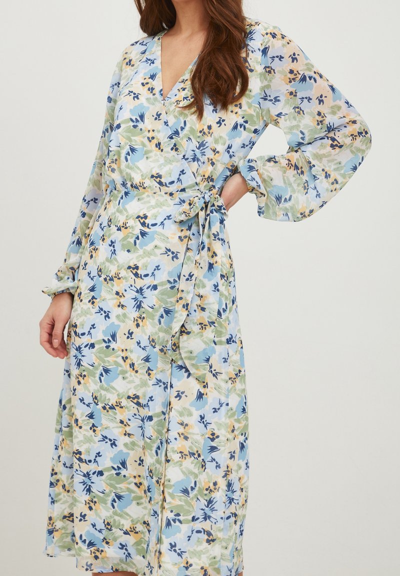 Femme portant une robe portefeuille à manches longues avec un motif floral bleu, vert et jaune et une ceinture nouée à la taille.