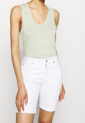 Vrouw draagt een lichtgroen mouwloos topje en witte knielange short, staand tegen een effen lichte achtergrond.