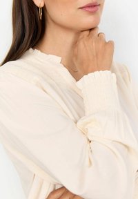 Cream blouse with long, puffed sleeves and ruffled cuffs. Features a textured bodice with smocked detailing along the neckline.