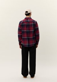 Mand iført rød og marineblå ternet skjorte, sorte bukser, beige kasket, stående med ryggen til mod en ensartet lys baggrund.