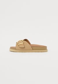 Sandales à glissière en suède tan avec un détail de boucle en ton or, semelle en caoutchouc plate et semelle intérieure rembourrée pour le confort. Design simple et minimaliste.