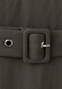 Gros plan d'une ceinture en tissu sombre avec une boucle rectangulaire recouverte de tissu et une épingle métallique la fixant à travers un œillet métallique.