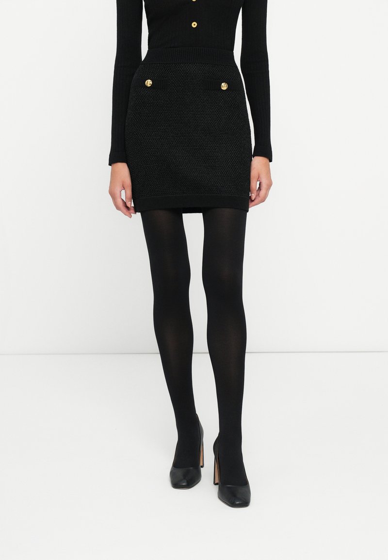 Jupe mini noire texturée avec deux boutons dorés sur les poches avant et une taille côtelée, associée à des collants noirs et des chaussures à talons.