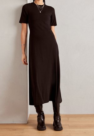 Schwarzes, kurzärmeliges Midikleid aus glattem, figurbetontem Stoff. Kombiniert mit kniehohen braunen Stiefeln mit Schnallen. Hintergrund: Holzboden.