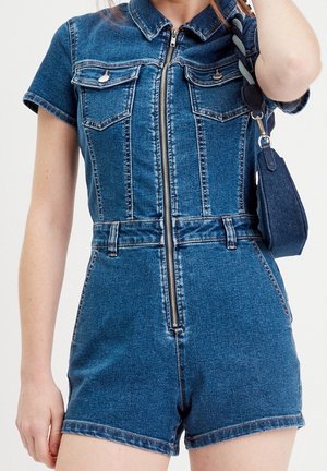 Femme portant une combinaison courte en jean à manches courtes avec une fermeture éclair à l'avant et des passants de ceinture, tenant un petit sac à main bleu foncé sur son épaule.