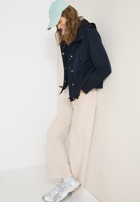 Femme appuyée contre un mur portant une casquette bleu clair, une veste à capuche bleu marine, un pantalon large beige et des baskets argentées, souriant en regardant vers le bas.