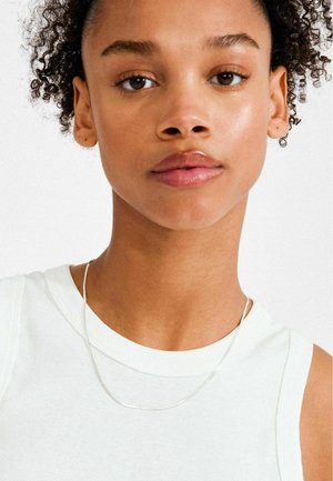 Srebrna verižica leži na belem brezrokavniku, poudarjajoč gladko površino in nežno ukrivljenost. Tonska barva kože je enakomerna z naravnim sijajem.