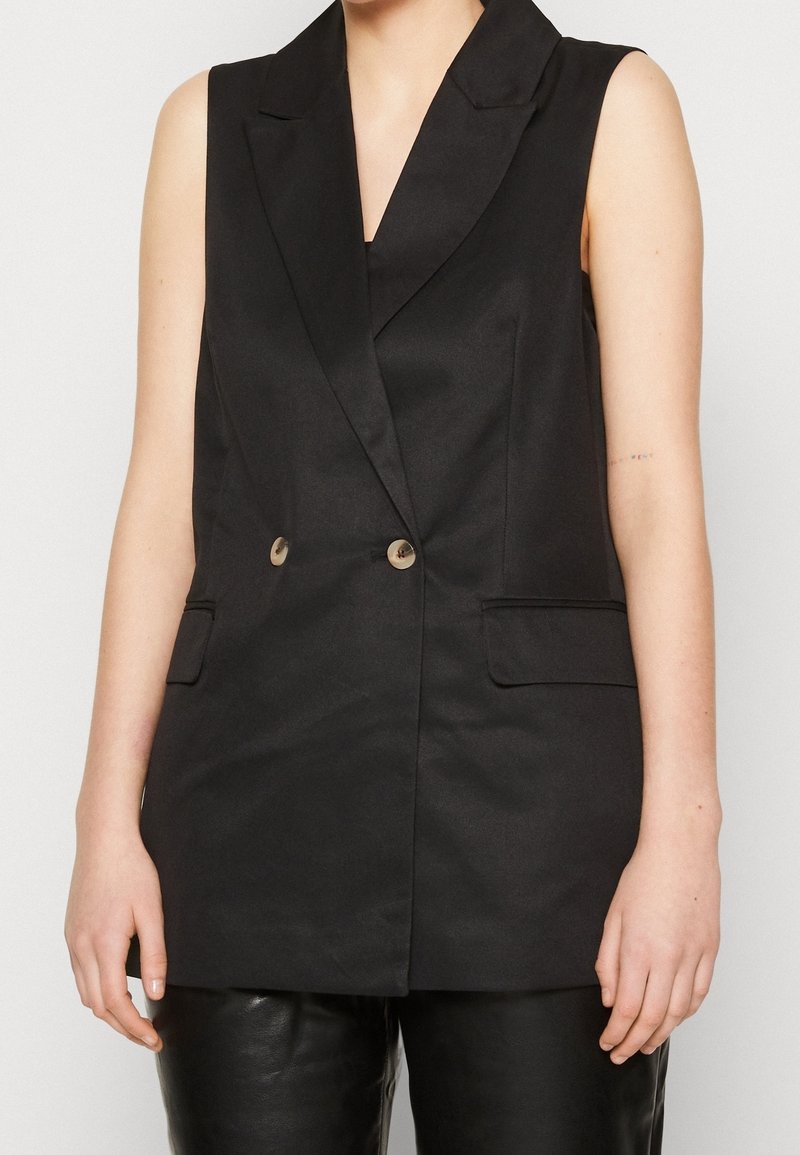 Blazer noir sans manches à double boutonnage avec revers et deux boutons, porté sur un pantalon en cuir noir par une personne debout.