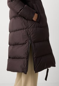 Manteau matelassé marron avec design en quilt, poches latérales zippées et fermeture éclair. Le tissu semble lisse avec une texture légèrement brillante.