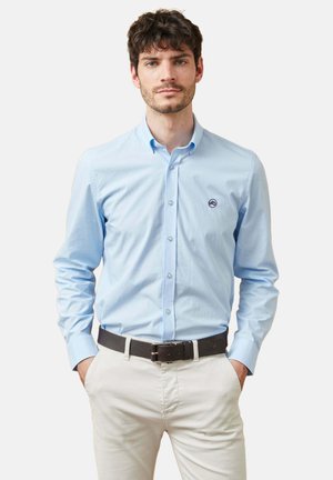 Hombre con el pelo corto y oscuro, que lleva una camisa celeste de botones, pantalones beige y un cinturón marrón oscuro, de pie con las manos en los bolsillos.