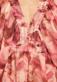 Diepe V-halsjurk met gerimpelde stoffen bandjes en een roze, rode en beige tie-dye patroon op lichtgewicht, vloeiend materiaal.