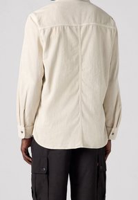 Jasno beżowa koszula z długim rękawem, z karczkiem z tyłu, szwami bocznymi i mankietami zapinanymi na guziki. Nosi się ją z czarnymi spodniami cargo z klapkami.