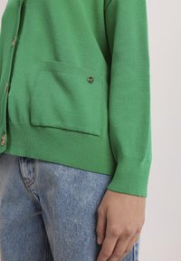 Zielony sweter robiony na drutach o teksturowym wykończeniu, z łatką na kieszeni i okrągłymi metalowymi guzikami. Noszony na jasnoniebieskich dżinsach.