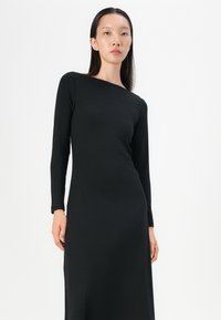 Robe noire à manches longues en coton côtelé avec un décolleté asymétrique et une silhouette ajustée. Texture douce avec un léger éclat.
