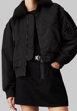 Personne portant une veste bomber noire courte avec col en peau lainée et une jupe noire avec un passant de ceinture marqué Calvin Klein.