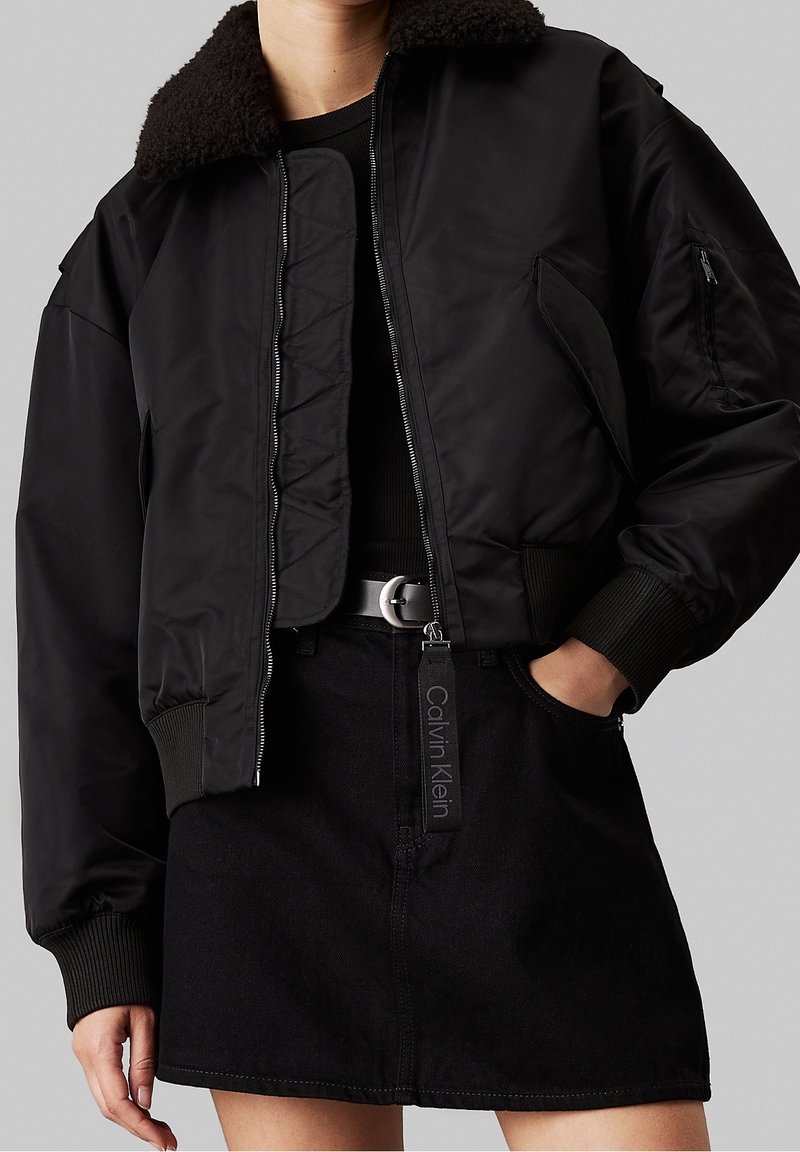 Personne portant une veste bomber noire courte avec col en peau lainée et une jupe noire avec un passant de ceinture marqué Calvin Klein.