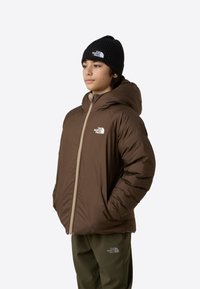 Jachetă izolată maro cu glugă, fermoar deschis și logo pe piept. Asortată cu pantaloni verde măsliniu, modelul poartă o căciulă neagră tricotată.
