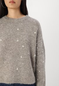 Pull en maille grise avec une texture douce et duveteuse, orné de perles éparpillées sur le devant et d'un col rond.
