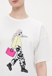 Hvit t-skjorte med en fargerik illustrasjon av en figur med langt hår, iført en gul topp, svarte blomstrete bukser, og som holder en rosa veske.