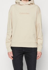Hoodie beige di Calvin Klein realizzata in un tessuto morbido, con tasca a marsupio frontale e polsini a coste. Il logo è stampato in una tonalità delicata.