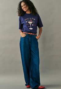 Mørkeblå t-shirt med en grafik af et cocktailglas, parret med brede mørke jeans i denim og et klart orange bælte. Røde sko fuldender outfittet.