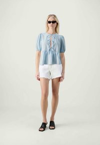 Blusa azul clara com mangas bufantes, detalhe de buraco e laços. Combinada com calções de ganga brancos e mules pretos de bico aberto.