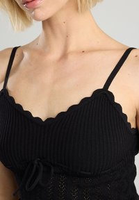 Black ribbed top with scalloped neckline, adjustable straps, and a fitted bodice featuring a textured pattern and drawstring detail at the waist.