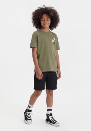 Göndör hajú gyermek, aki olívazöld mintás pólót, fekete rövidnadrágot, csíkos zoknit és fekete magas szárú tornacipőt visel, és fehér háttér előtt áll.