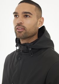 Chaqueta impermeable negra con un alto cuello, capucha ajustada y cierres de botón. Textura suave y diseño minimalista.