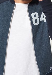 Marinblå bomberjacka med en grå dragkedja, med vit "84" på bröstet. Lagd över en vit t-shirt med rund hals.