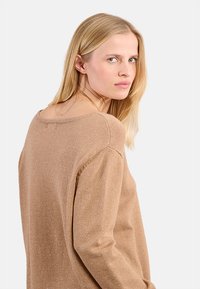 Donna con lunghi capelli biondi che indossa un maglione di lana beige, mentre guarda oltre la spalla su uno sfondo chiaro e semplice.