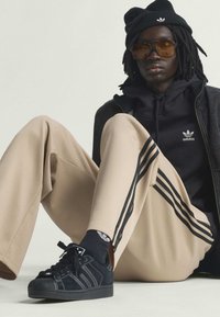 Osoba v čiernej čiapke, tmavých slnečných okuliaroch, čiernej mikine Adidas, béžových teplákoch Adidas s čiernymi pruhmi, čiernych ponožkách a čiernych teniskách sediaca na zemi.