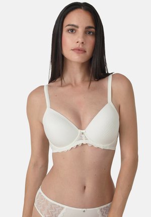 Junge Frau mit langen dunklen Haaren, die einen weißen gepolsterten BH und passende Spitzenunterwäsche trägt, stehend vor einem schlichten hellen Hintergrund.