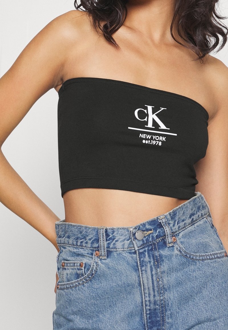 Černý top bez ramínek s cropped střihem a bílým logem "cK NEW YORK est. 1978", spárovaný s džínovými kraťasy s vysokým pasem, které mají modrý prát a knoflíkové detaily.